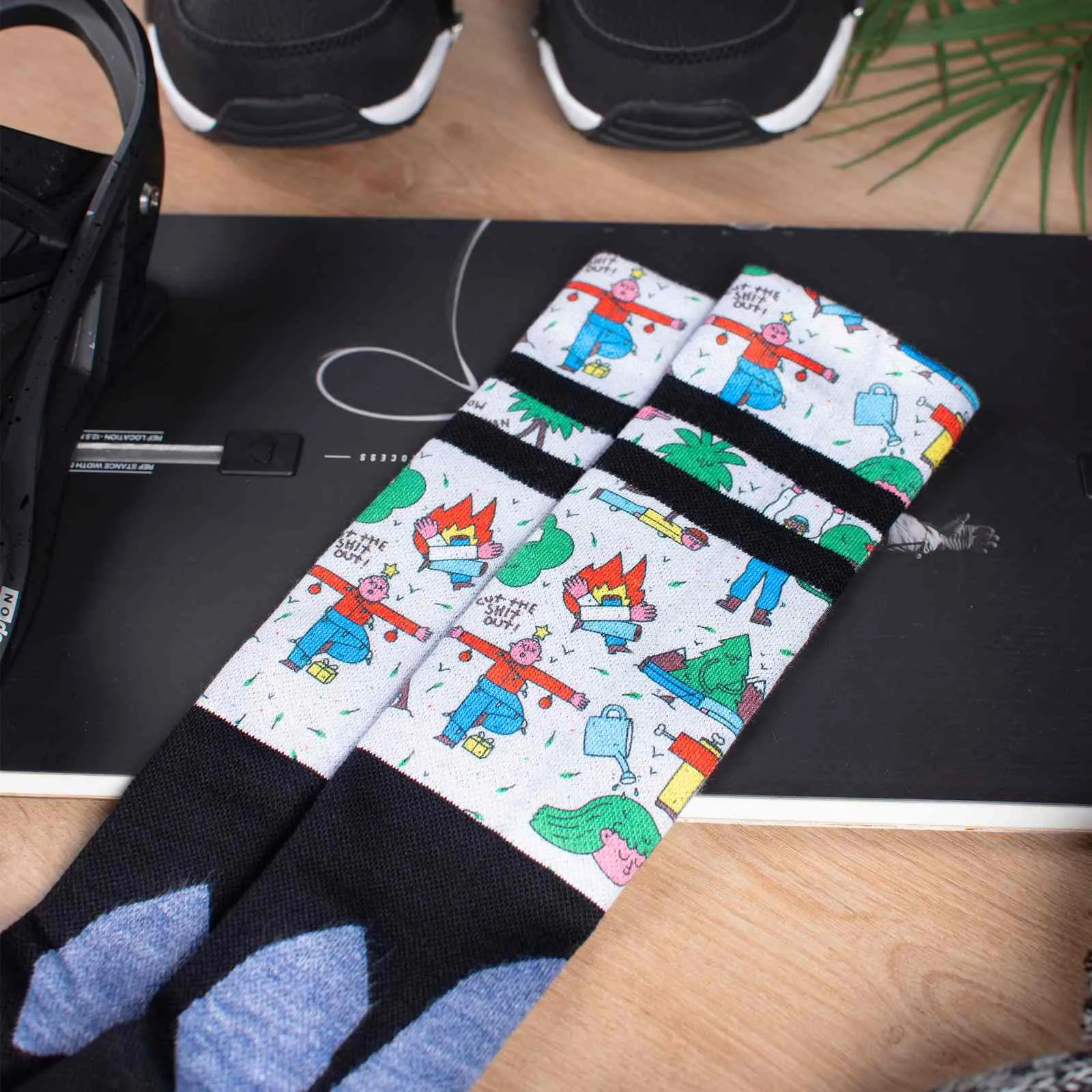 Kniehohe Snow Socks auf Snowboard, Motiv mit Comicfiguren, schwarz-weiß gestreift, dazu Snowboard Boots und ein Blatt.