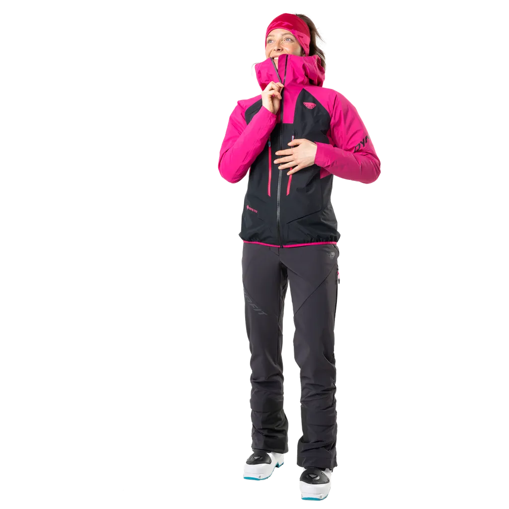 Frau in Dynafit TLT Touring DST Hose, schwarz, pinke Jacke, Skischuhe, Blick nach oben.