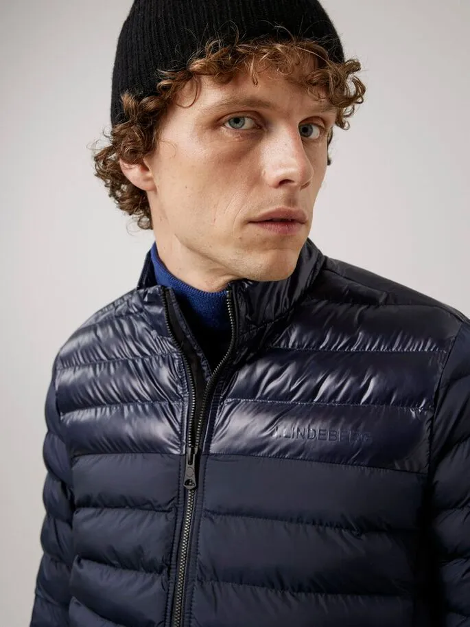 Mann mit Mütze trägt blaue, wattierte Rick Light Jacket mit Reißverschluss und kleinem J.Lindeberg Schriftzug.