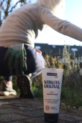 Eine Tube Handbalsam steht im Vordergrund, im Hintergrund eine Person mit Gartenhandschuhen kniet im Garten.