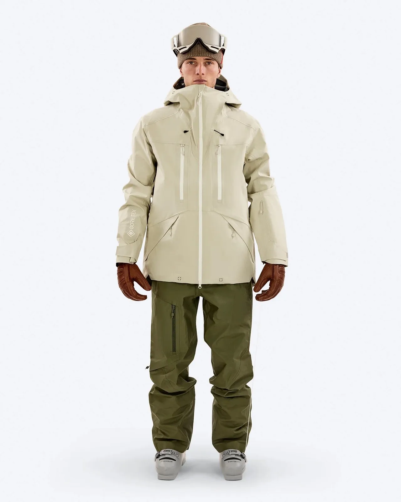 Eine Person in Skikleidung steht vor einem weißen Hintergrund. Sie trägt eine beigefarbene Jacke, eine grüne Hose, braune Handschuhe und eine Skibrille.