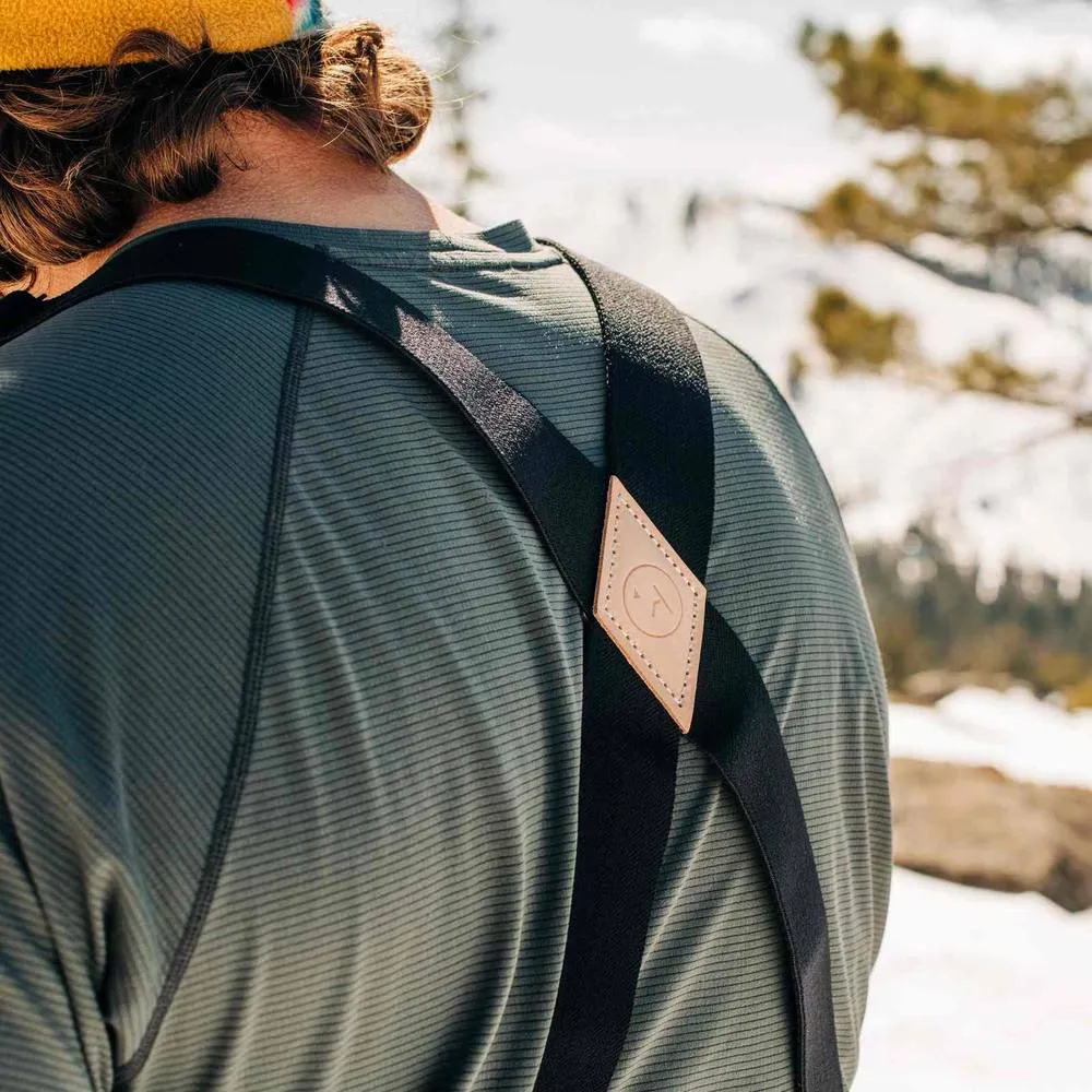 Schwarze Hosenträger mit Lederdetail auf einem Mann mit gestreiftem Shirt vor verschwommenem Hintergrund.