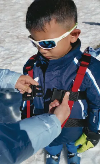 Kind mit Skibrille und Skitrainer-Gurt, der von einer Person geschlossen wird.