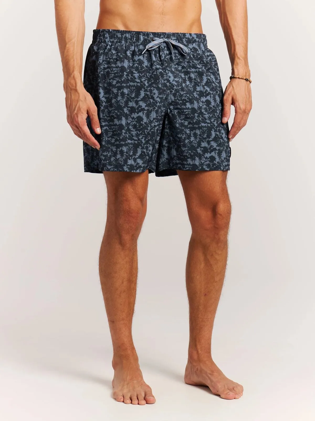 Ein Mann trägt Badeshorts mit Palmenmuster, stehend vor einem weißen Hintergrund.