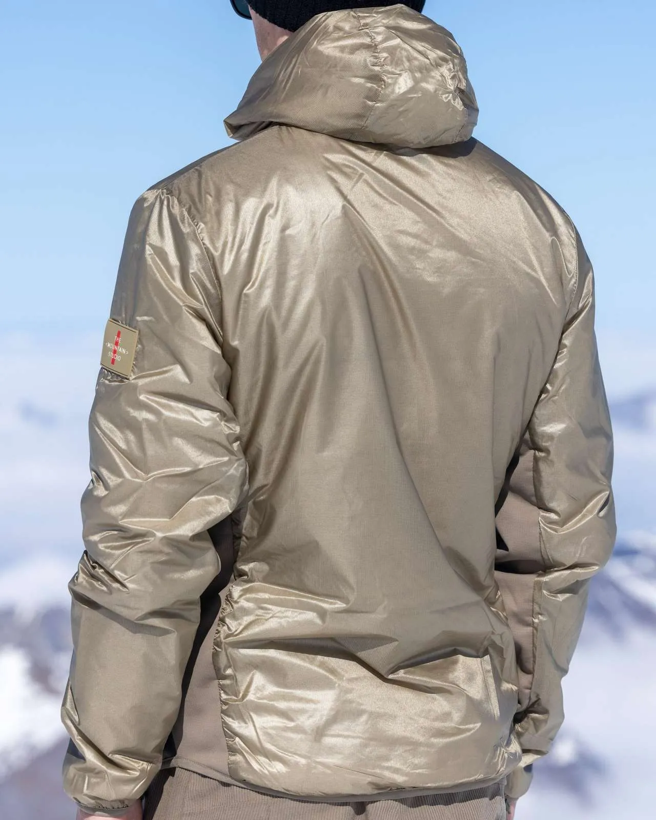 Rückenansicht einer Person mit goldener Daunenjacke vor verschneiter Bergkulisse und blauem Himmel.