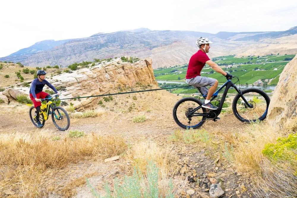 Zwei Personen fahren E-Bikes, verbunden durch ein Abschleppseil, in hügeliger Landschaft.