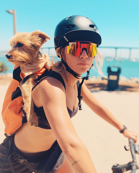 Frau mit Helm und polarisierter Sonnenbrille, trägt Hund im Rucksack.