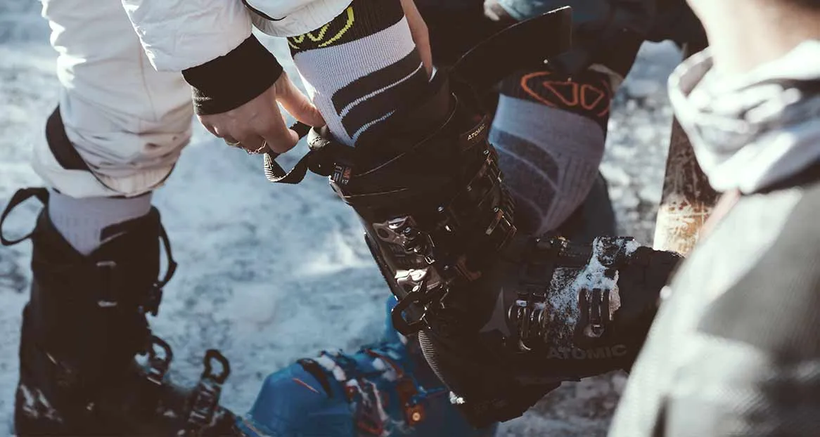 Nahaufnahme von Skischuhen, die auf Schnee stehen. Eine Person befestigt einen Skischuh. Skisocken sind sichtbar.
