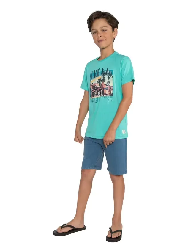 Ein Junge steht barfuß da und trägt ein türkisfarbenes T-Shirt mit einem Aufdruck eines Autos, Palmen und der Aufschrift "Surf & Co." sowie blaue Shorts. Er hat braune Haare und trägt schwarze Flip-Flops.