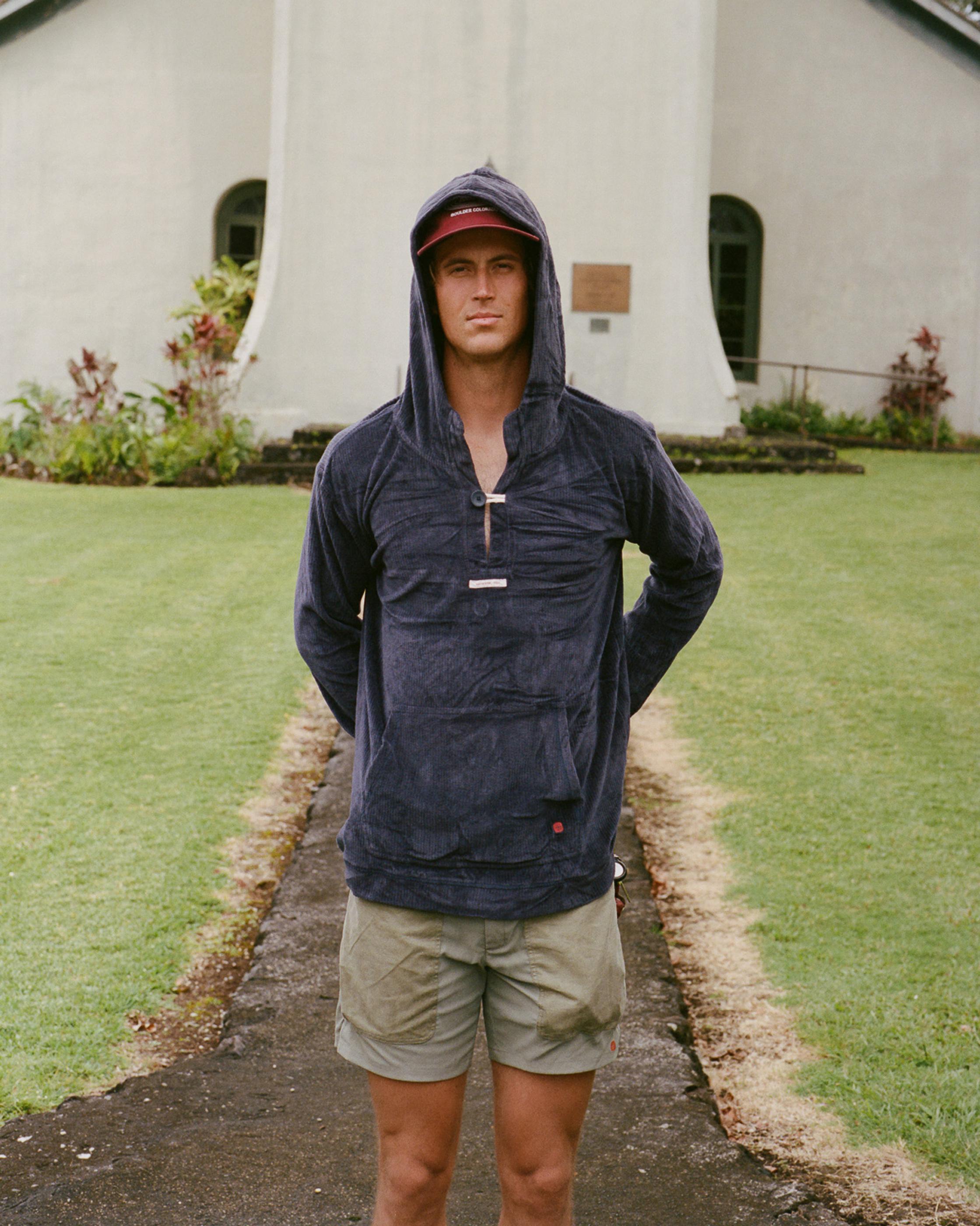 Ein Mann mit Mütze und Kapuzenpulli steht vor einem Gebäude mit Rasenfläche.