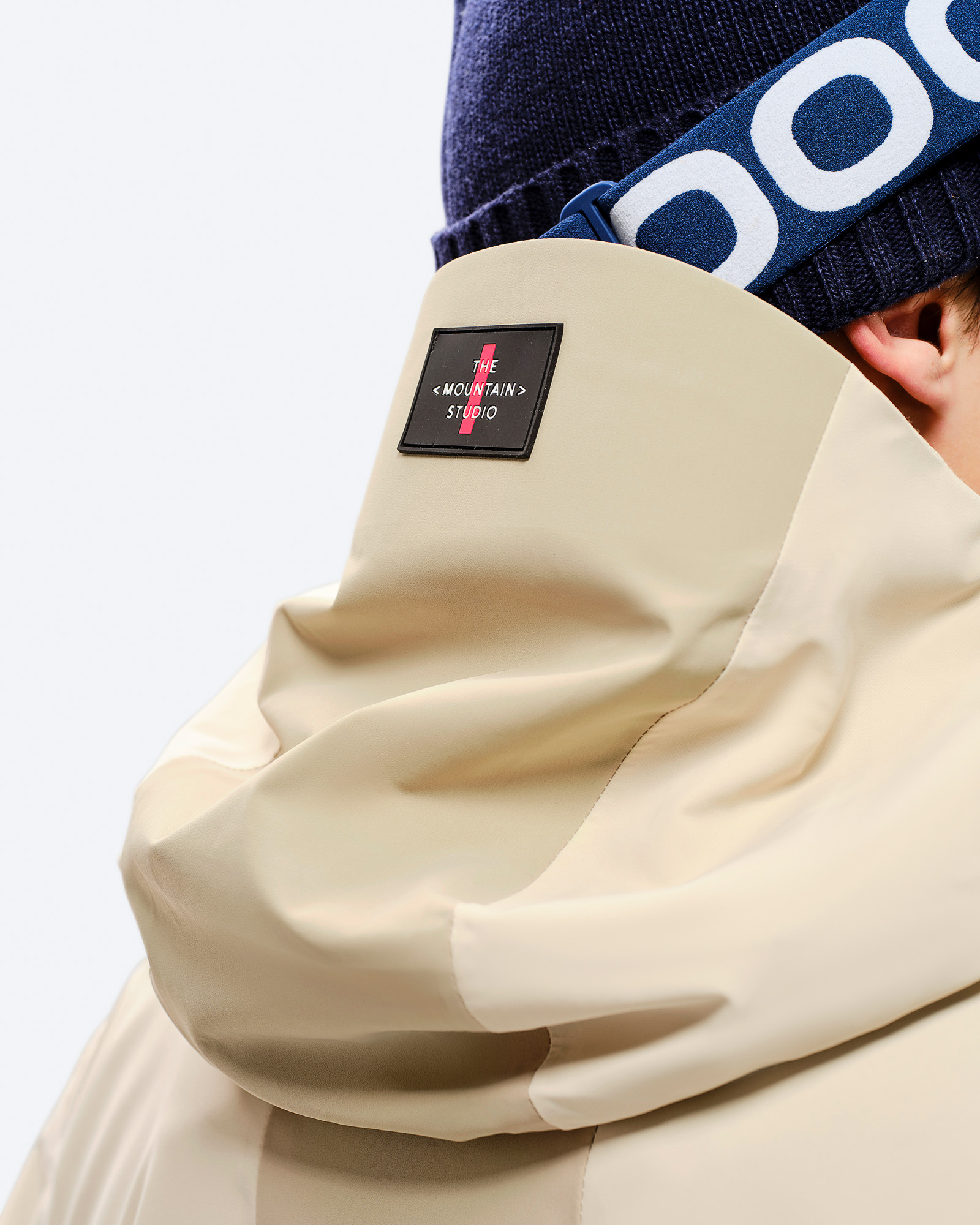 Nahaufnahme der Kapuze einer beigen Z-5 3L Hybrid Shell Jacke, mit Mütze und Skibrille im Hintergrund.