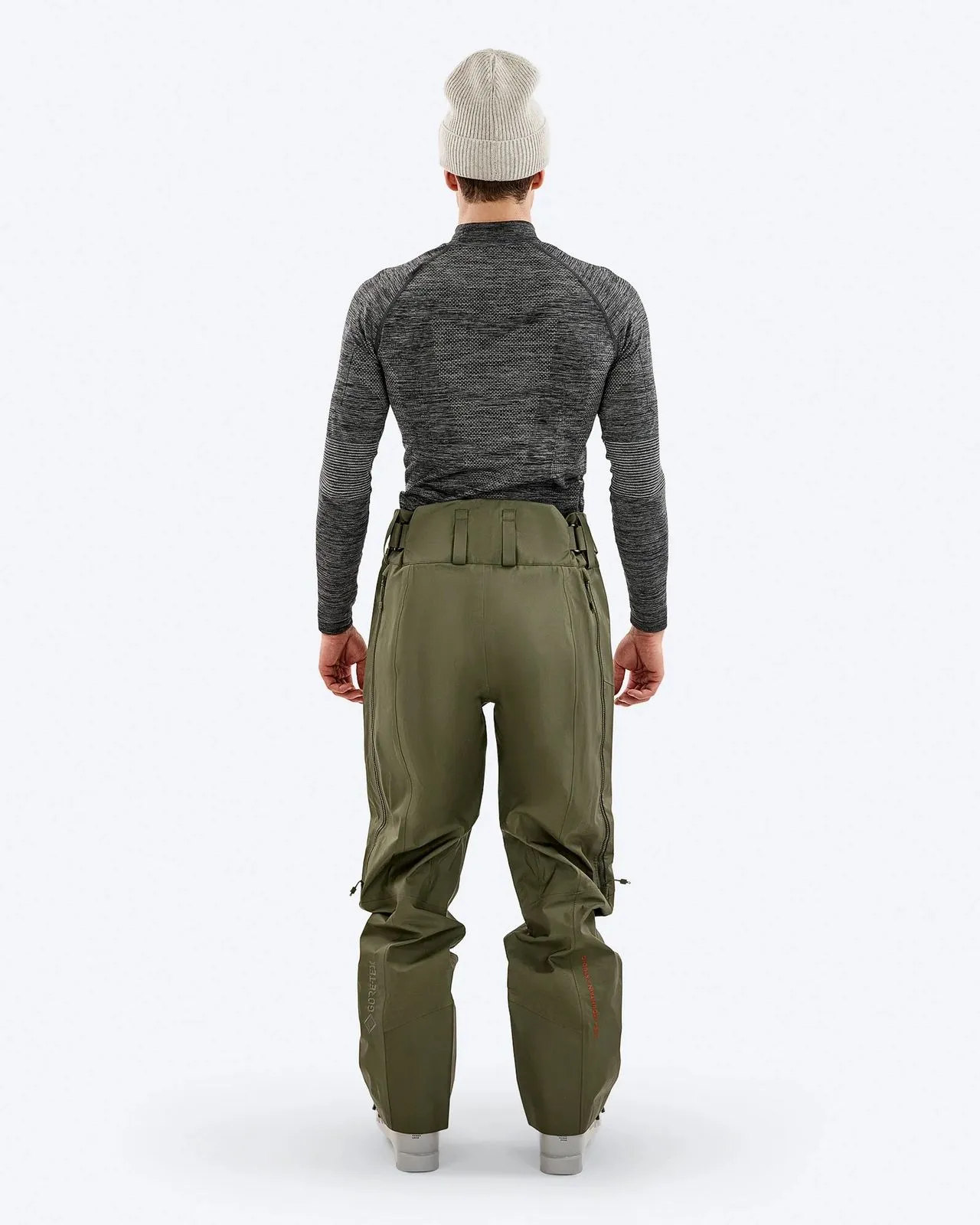Ein Mann von hinten, trägt eine graue Mütze, ein graues Langarmshirt und olivgrüne Hose. Die Hose hat Reißverschlüsse an den Seiten.