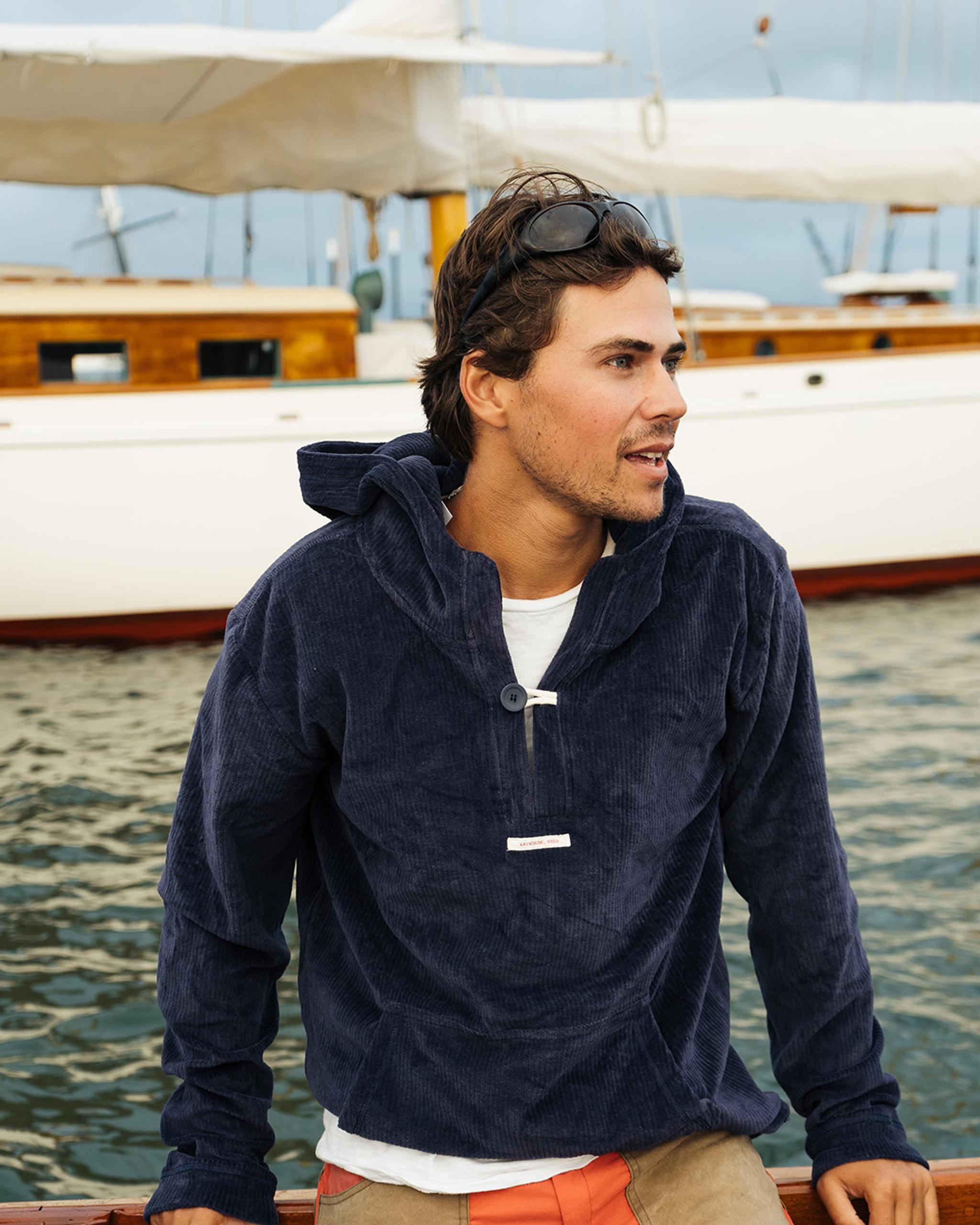 Ein Mann in einem blauen Cordhoodie lehnt an einer Yachtreling. Er trägt eine Sonnenbrille auf dem Kopf und schaut nach rechts.