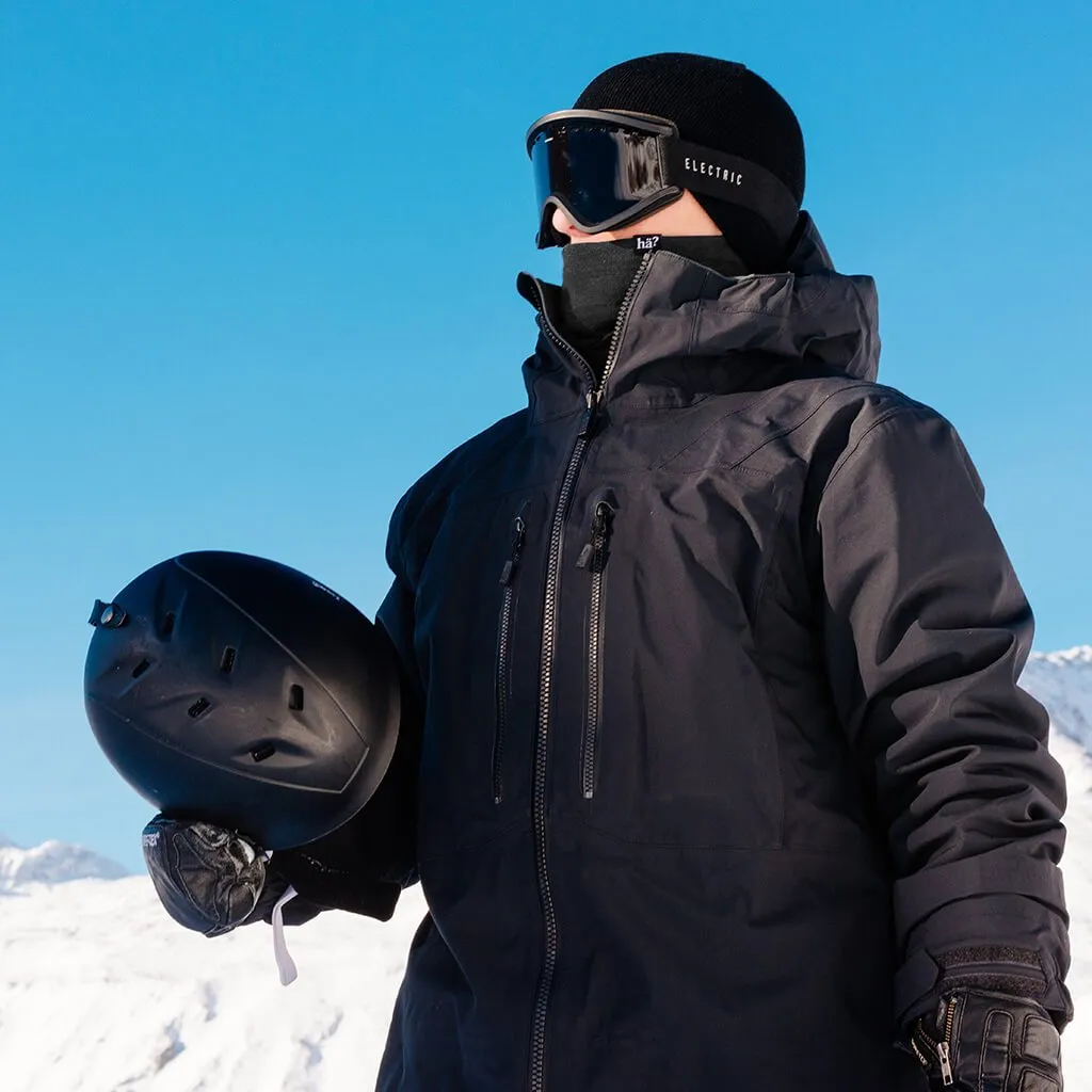 Ein Mann mit schwarzer Jacke, Beanie, Brille und Neckwarmer hält einen Helm in einer Winterlandschaft.