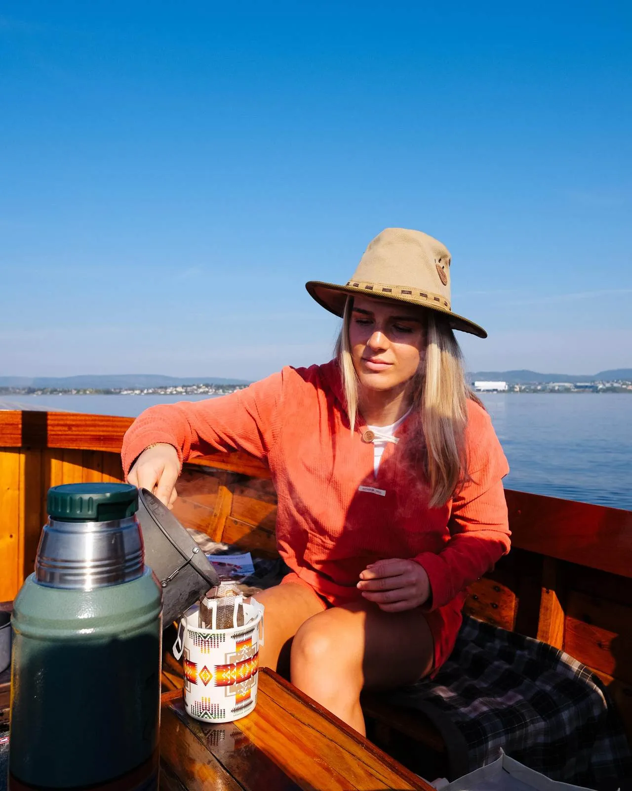 Eine Frau mit Hut gießt dampfenden Kaffee in eine Tasse auf einem Holzboot mit blauem Himmel und Wasser im Hintergrund.