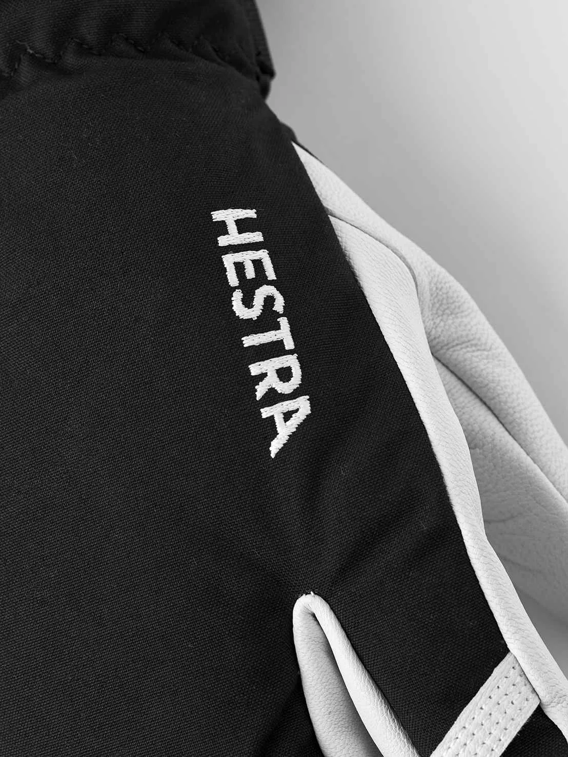 Nahaufnahme eines schwarzen Hestra-Handschuhs mit weißem Lederbesatz und aufgesticktem Logo.