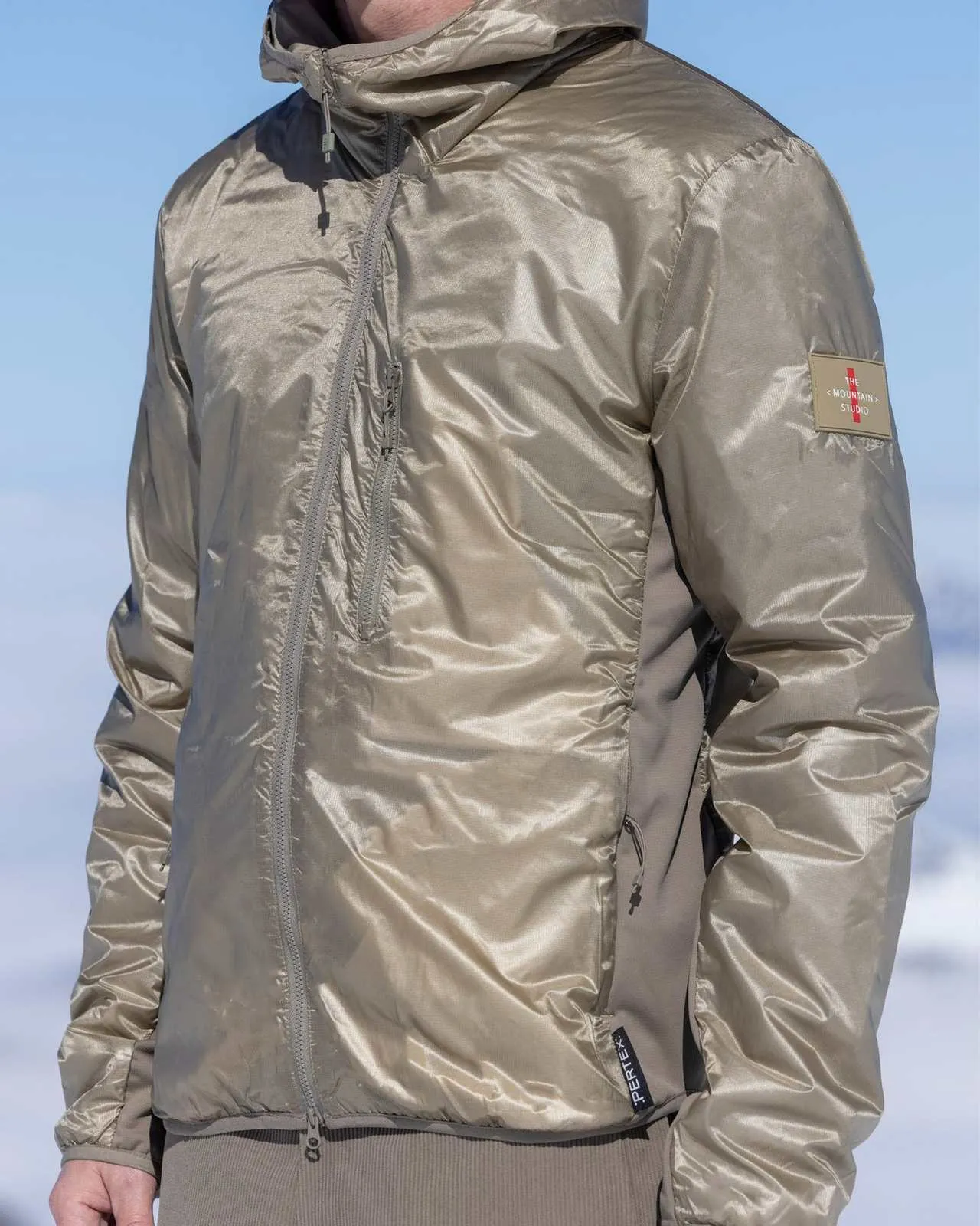 Mann in glänzender, beiger Jacke mit Kapuze vor verschneitem Hintergrund. Details: Reißverschlüsse, Ärmelpatch, reflektierendes Material.