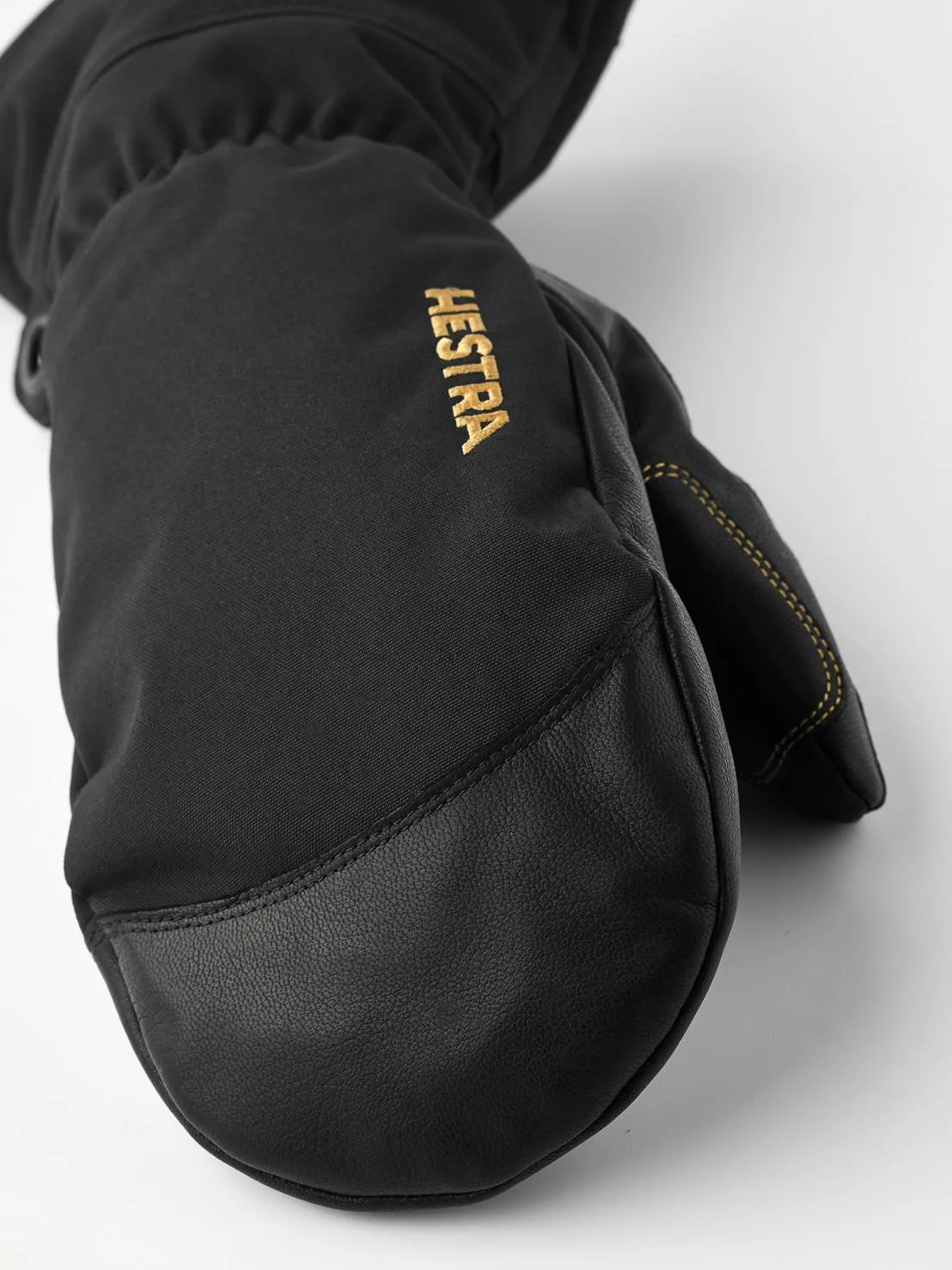 Detailaufnahme eines schwarzen Hestra Fäustlings aus Leder und Stoff, mit dem goldfarbenen Hestra-Logo auf dem Handrücken.