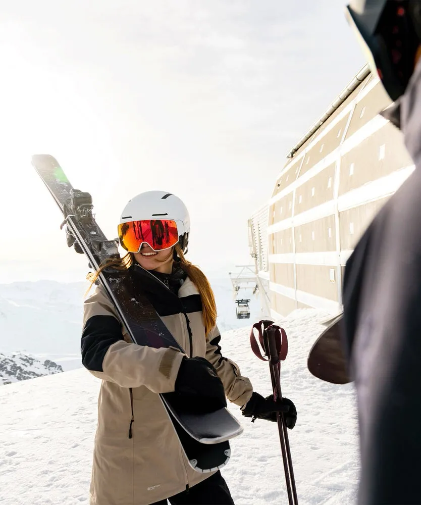 Skifahrerin mit weißem Helm (Driver Prime Sigma Photo Mips) und Skiern auf der Schulter im Schnee. Sonnenlicht und Bergpanorama im Hintergrund.