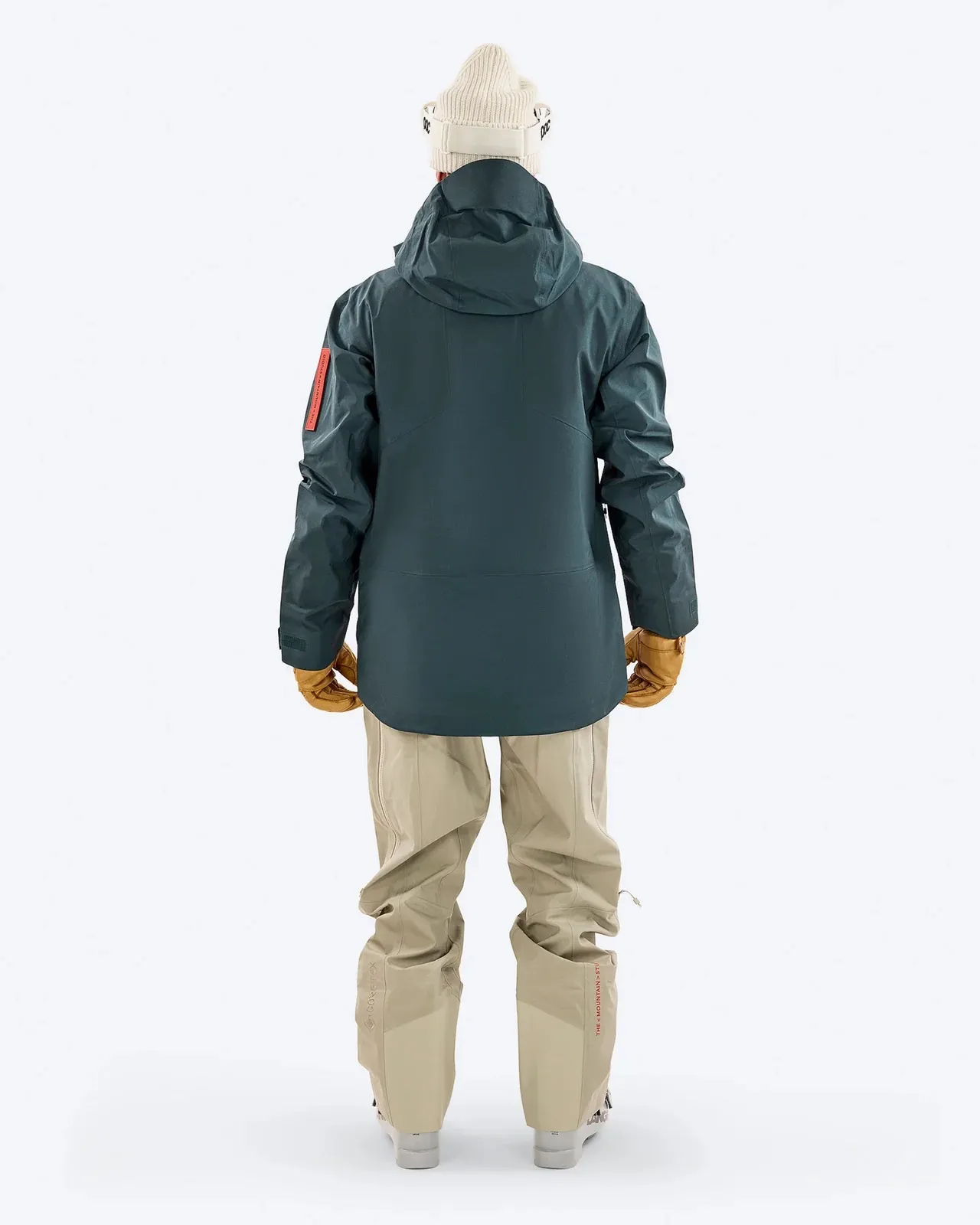 Person von hinten, mit grüner Jacke, beigen Hosen, Handschuhen und Mütze vor weißem Hintergrund.