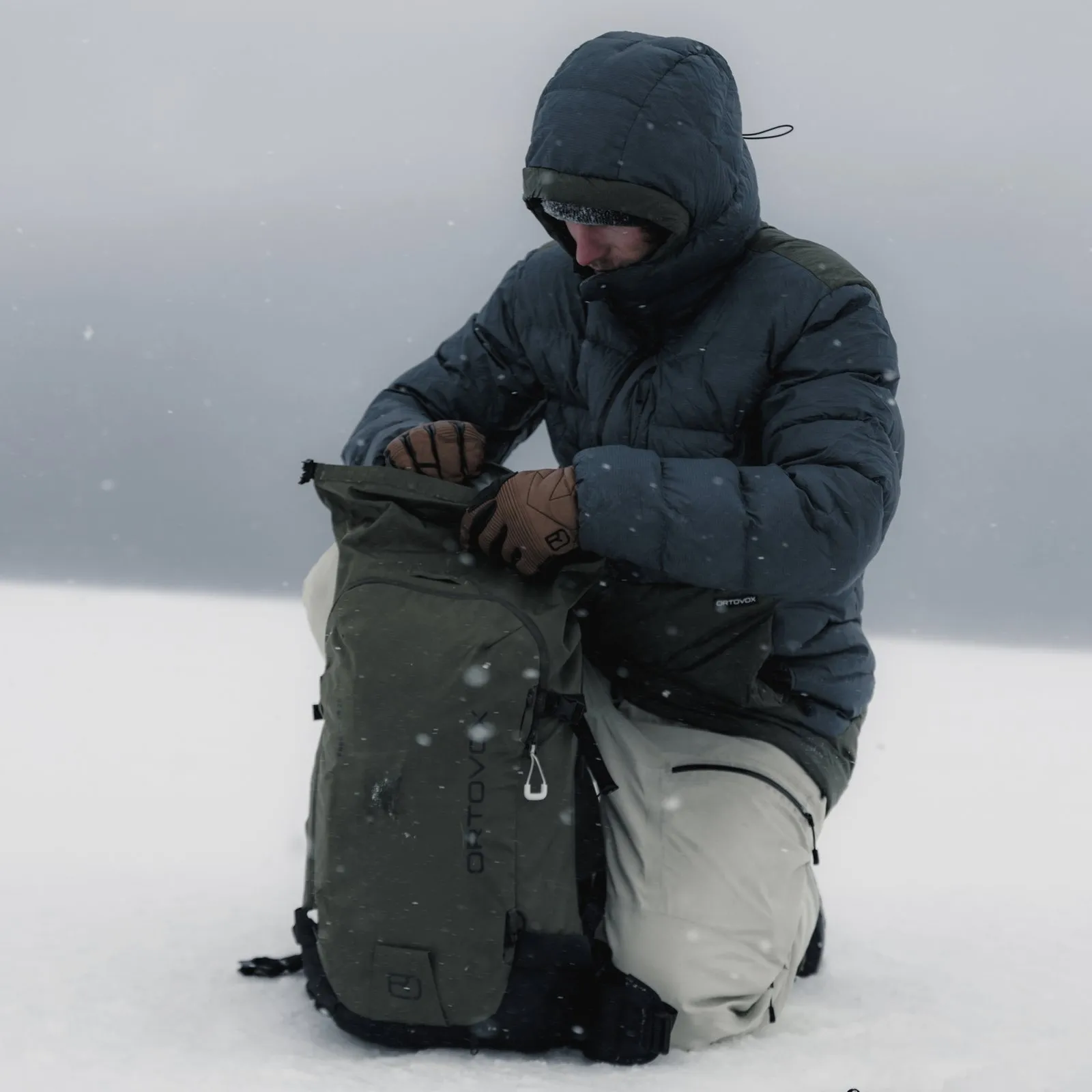 Ein Mann kniet im Schnee, er trägt eine Daunenjacke und öffnet einen olivgrünen Rucksack. Es schneit leicht.