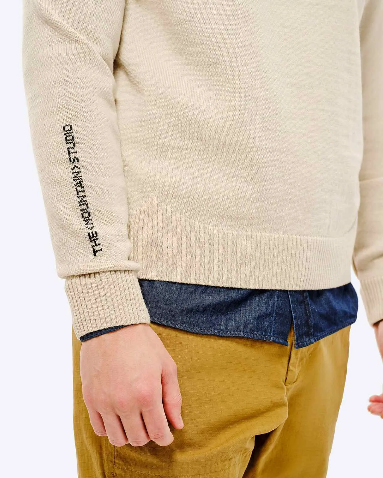 Detailansicht vom St. Moritz Ski Sweater, beige, mit Schriftzug auf dem Ärmel, über einem blauen Hemd und beigen Hosen.