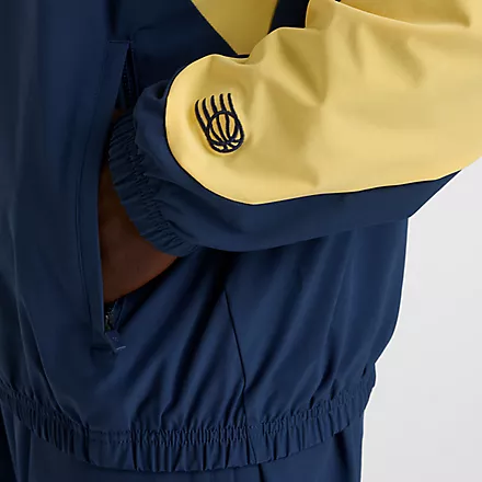 Detailaufnahme der New Balance In Game Woven Track Jacket in Navy/Gelb. Sichtbar: Ärmel-Logo, Seitentasche mit Reißverschluss und elastischer Saum.