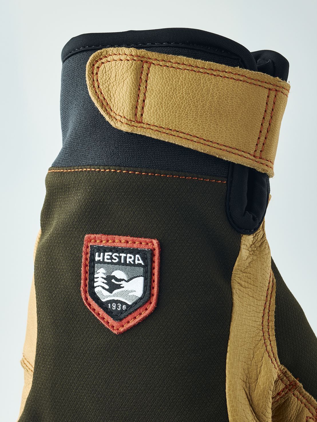 Braune Lederhandschuhe Detailaufnahme eines Handschuhs mit braunem Obermaterial, Lederdetails und "Hestra 1936" Logo. Der Handschuh hat eine Stulpe mit Klettverschluss.
