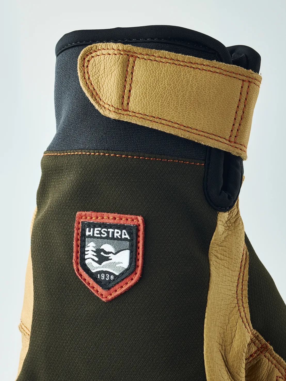 Detailaufnahme eines Handschuhs mit braunem Obermaterial, Lederdetails und "Hestra 1936" Logo. Der Handschuh hat eine Stulpe mit Klettverschluss.