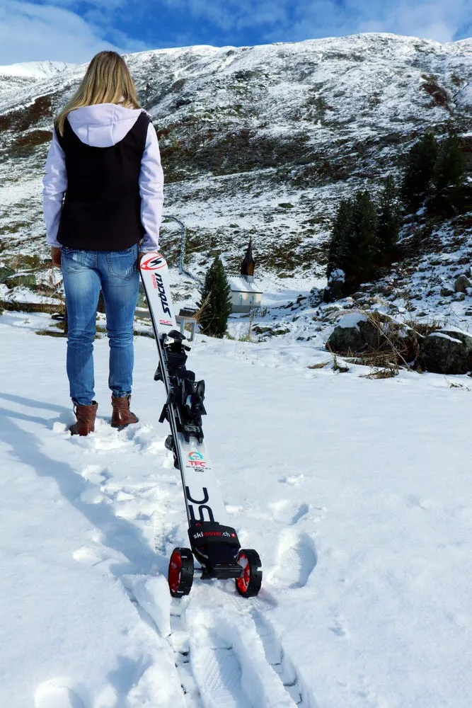 Frau mit Skiern auf Schnee, ausgestattet mit rotem Skimover Traveller Pro mit Rädern. Im Hintergrund ist ein Berg und blauer Himmel sichtbar.