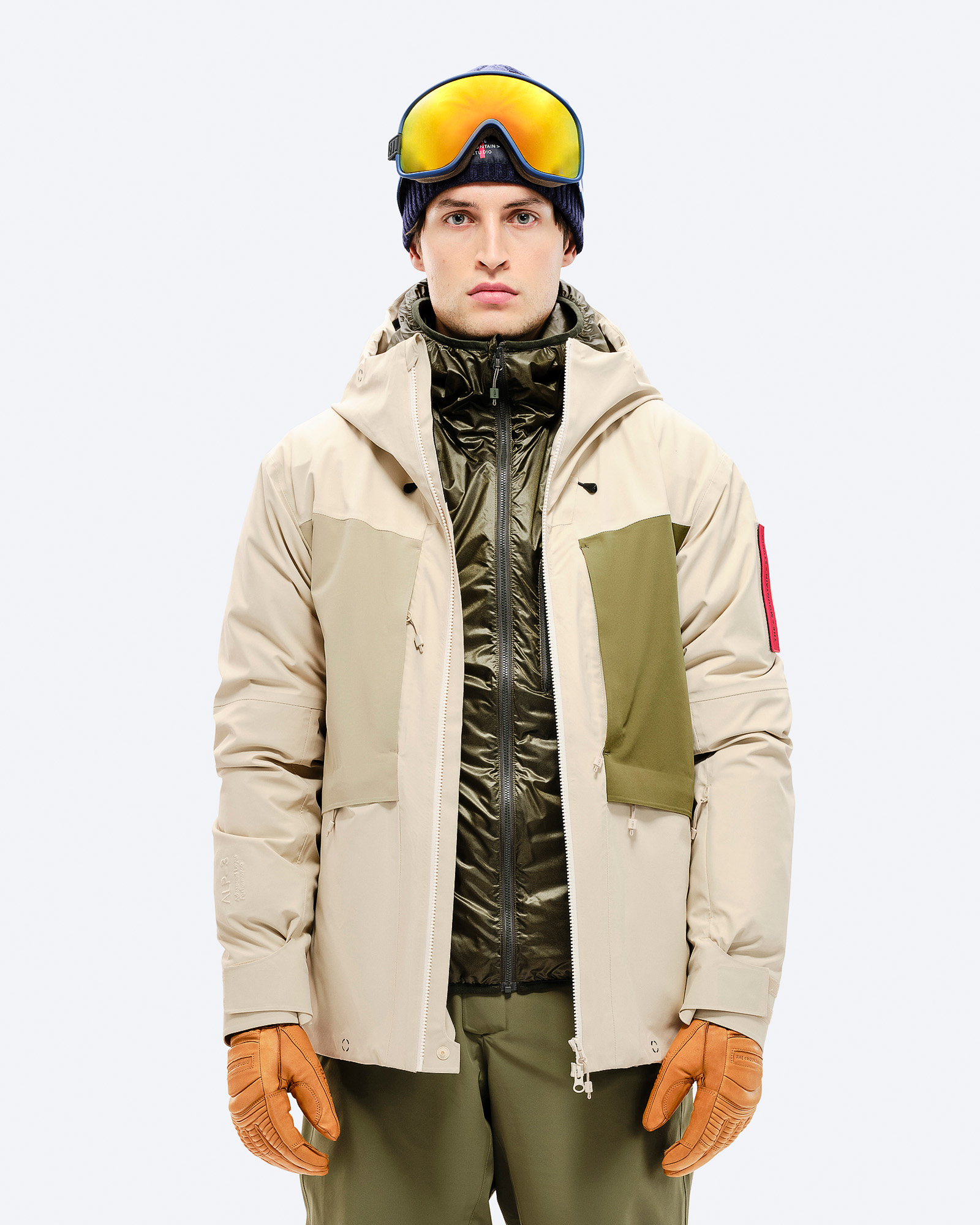 Mann mit Skibrille und Mütze, trägt beige 3L Hybrid Shell Jacke mit grünen Akzenten und Lederhandschuhen.