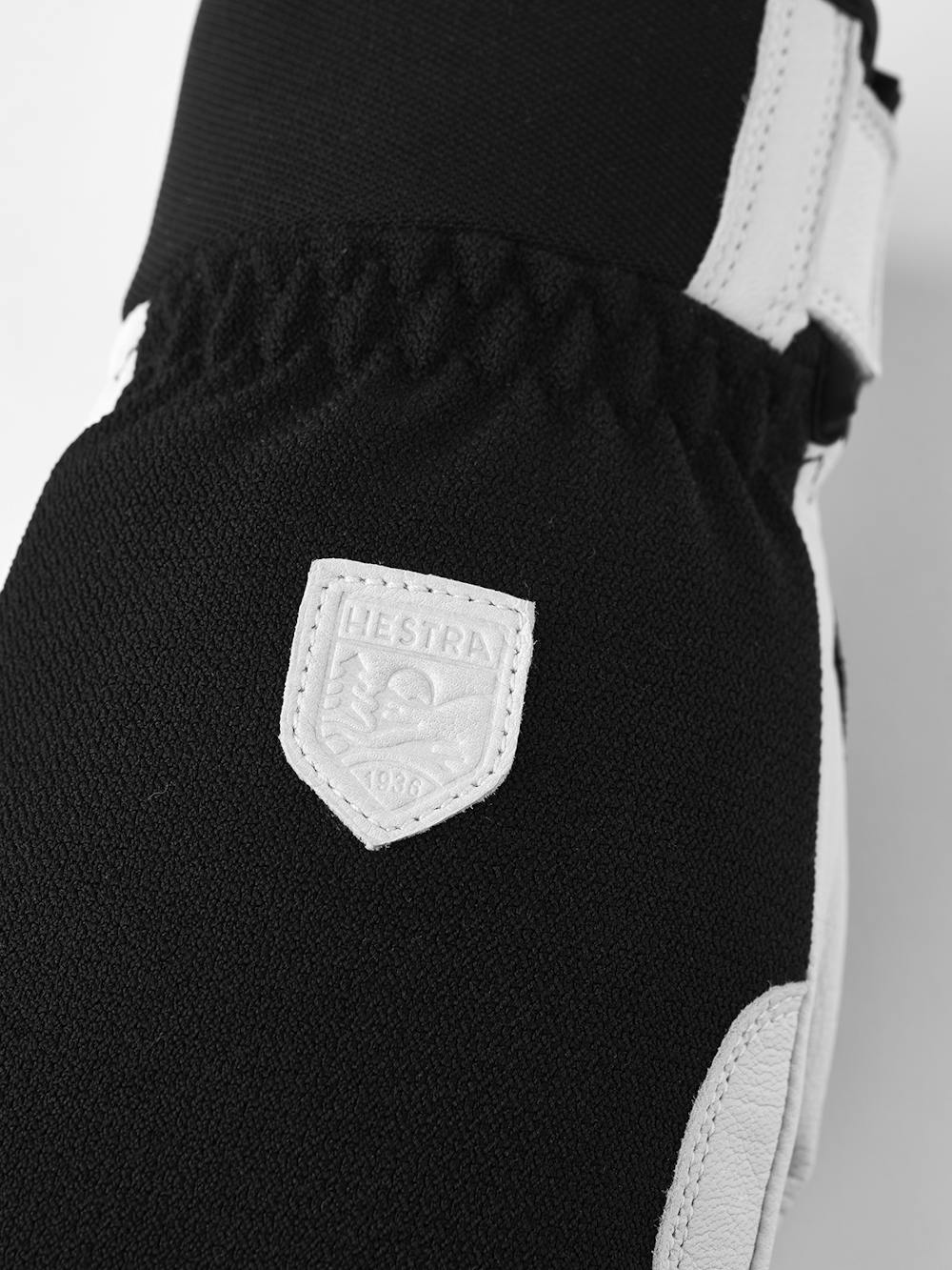 Nahaufnahme eines schwarzen Handschuhs mit weißem Lederakzent und einem Hestra-Logo.