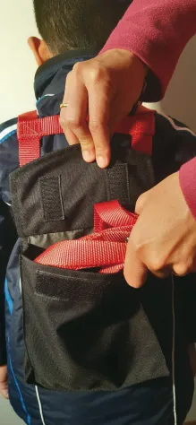 Kind mit Skitrainer: Rote Leine wird in eine schwarze Tasche auf dem Rücken verstaut. Elternhände falten die Tasche zu.