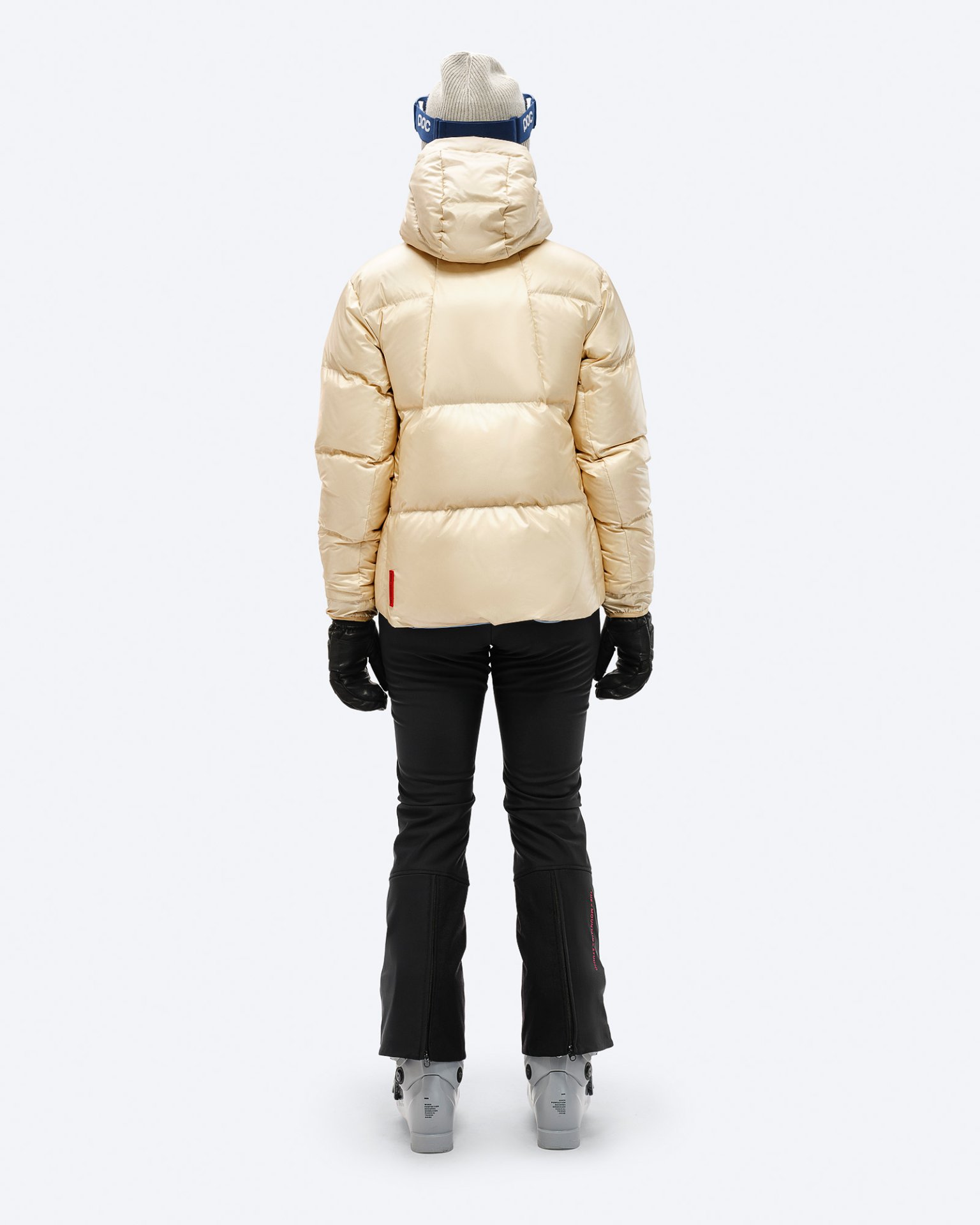 Rückansicht: Beige Daunenjacke mit Kapuze, schwarze Skihose, Handschuhe und Skischuhe, vor einem weißen Hintergrund.