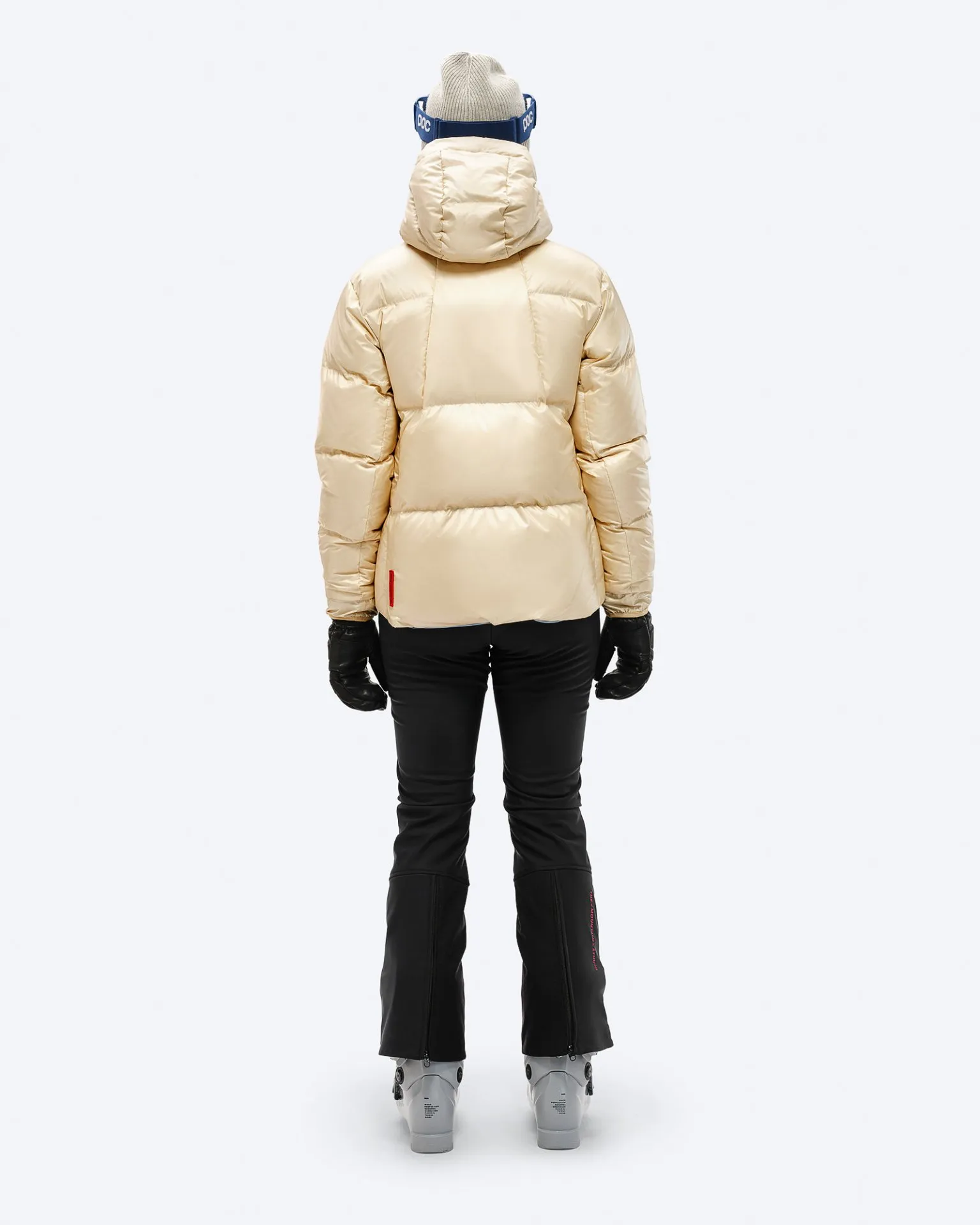 Rückansicht: Beige Daunenjacke mit Kapuze, schwarze Skihose, Handschuhe und Skischuhe, vor einem weißen Hintergrund.