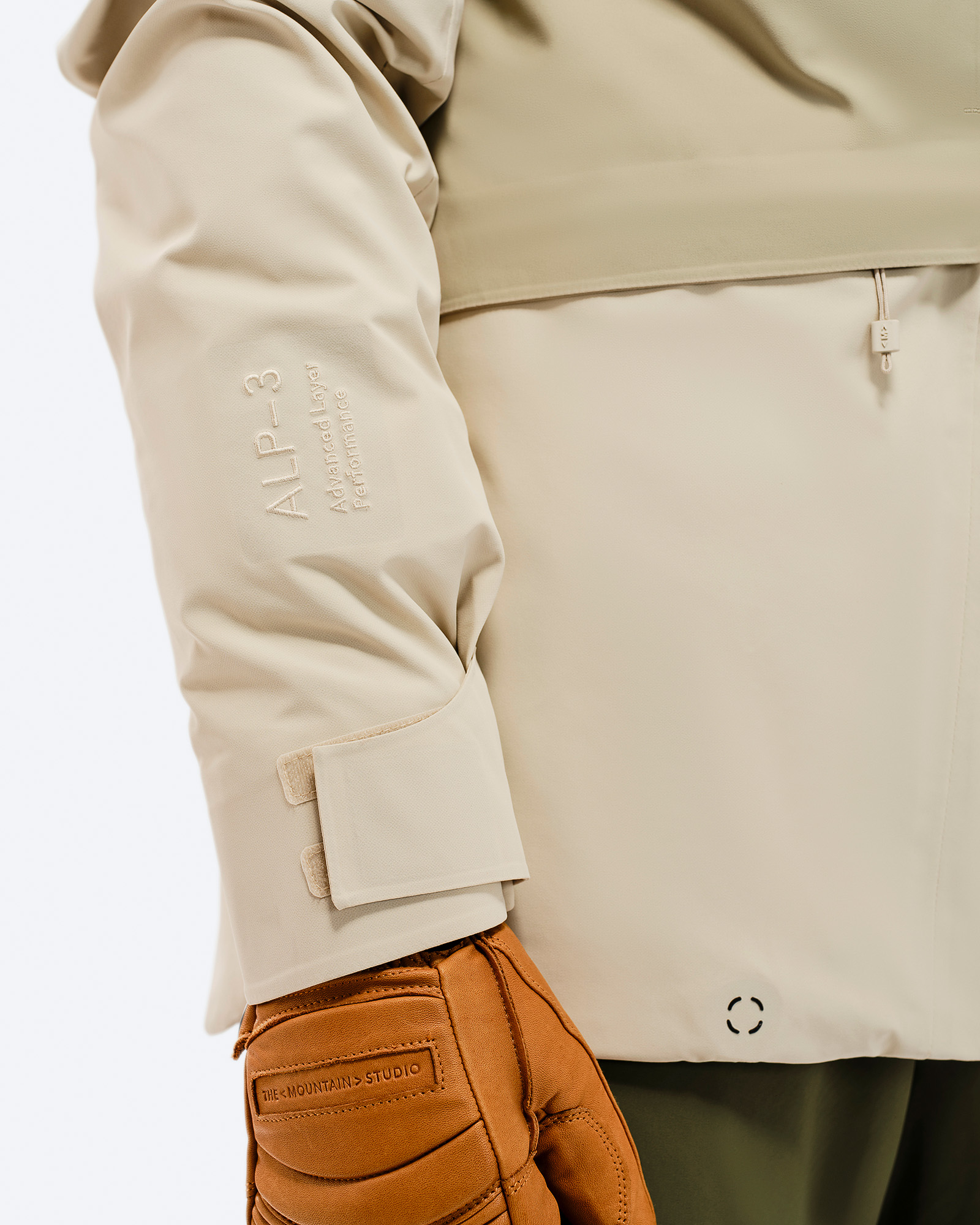 Detailaufnahme der beigen Z-5 3L Hybrid Shell Jacket mit ALP-3 Logo am Ärmel und braunem Lederhandschuh.