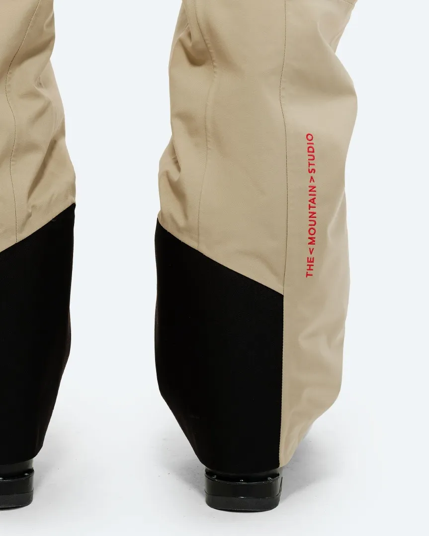 Detailaufnahme: Untere Hälfte der P-7 Ride Insulated Pant in Beige mit schwarzem, diagonalen Farbblock und rotem Markenlogo.