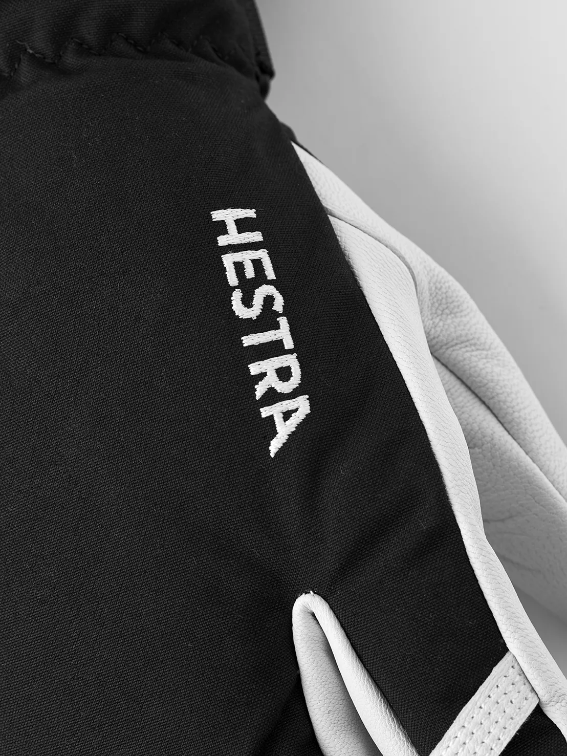Nahaufnahme eines schwarzen Hestra-Handschuhs mit weißem Lederbesatz und aufgesticktem Logo.