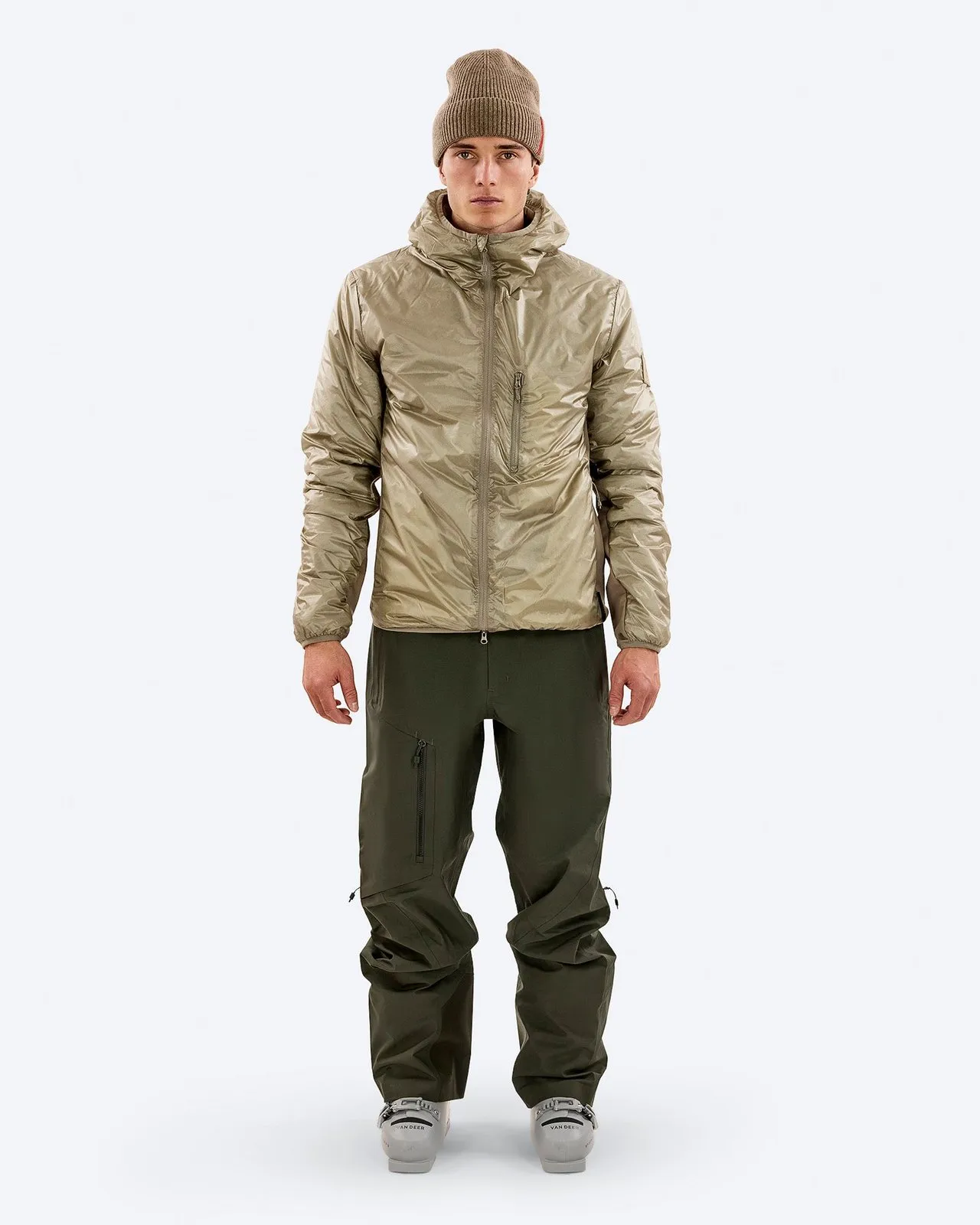 Mann in Outdoor-Kleidung: Braune Mütze, schimmernde Jacke mit Kapuze, olivgrüne Hose und Skischuhe vor weißem Hintergrund.