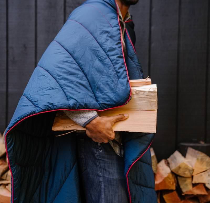 Mann mit Rumpl Puffy Blanket Deepwater, trägt Holzscheite vor Holzwand.