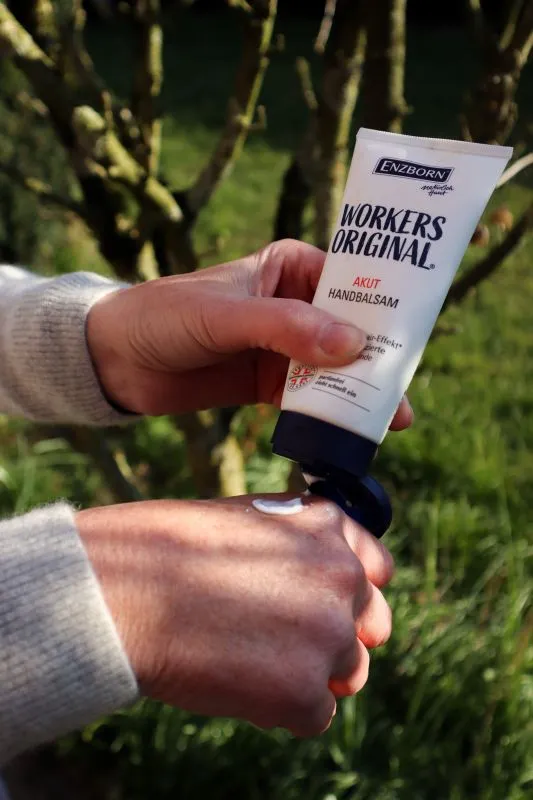 Eine Person trägt Enzborn Workers Original Handbalsam auf den Handrücken auf. Im Hintergrund sind Bäume und Gras zu sehen.