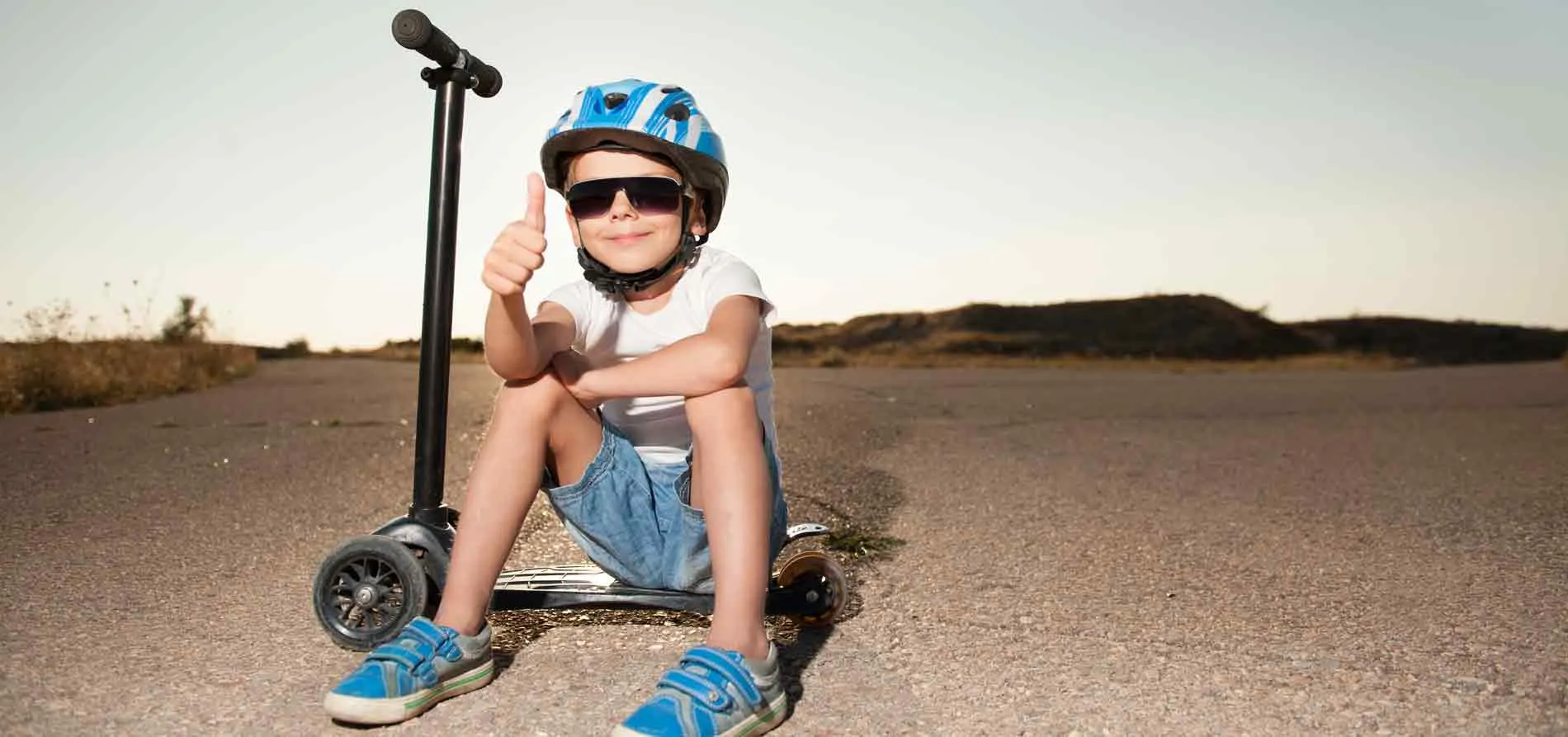 Junge mit Sonnenbrille und Helm sitzt auf Roller und zeigt Daumen hoch.