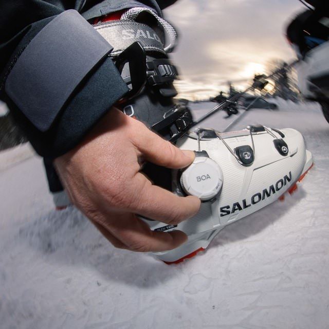 Nahaufnahme: Hand bedient BOA-Drehverschluss am weißen Salomon S/PRO SUPRA BOA 120 Skischuh im Schnee.