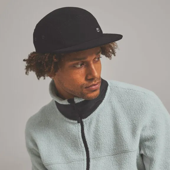 Ein Mann mit lockigem Haar trägt eine schwarze Kappe und eine hellblaue Fleecejacke. Er schaut zur Seite.