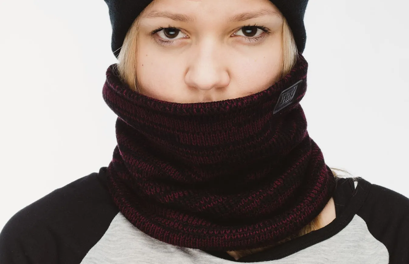 Porträt einer Person mit bordeauxfarbenem Melange-Neckwarmer, der bis zur Nase hochgezogen ist, und einer schwarzen Mütze.