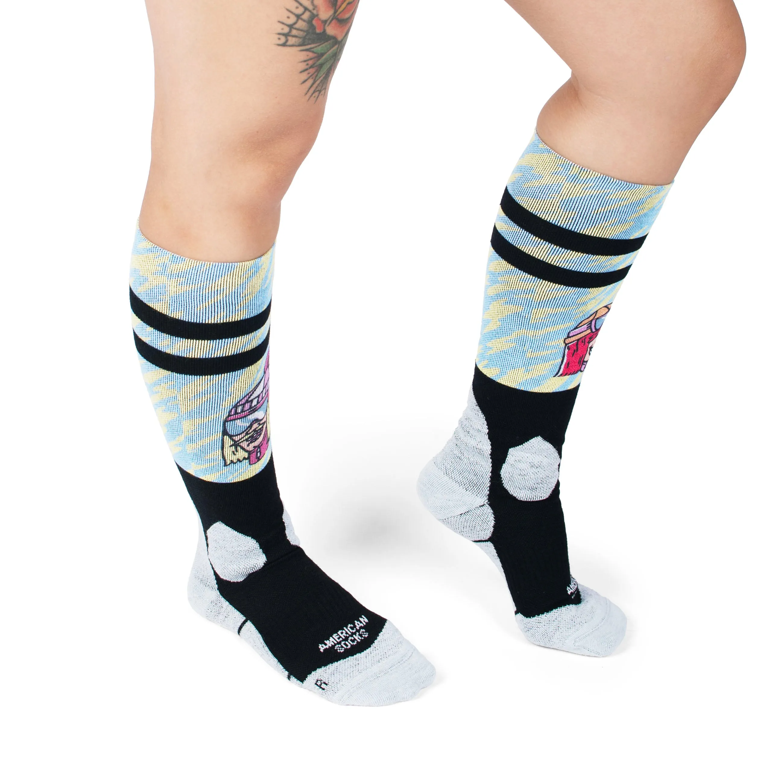 Beine mit gemusterten Slope Dope Snow Socks, schwarz abgesetzt mit American Socks Logo. Muster mit Strand Motiv.