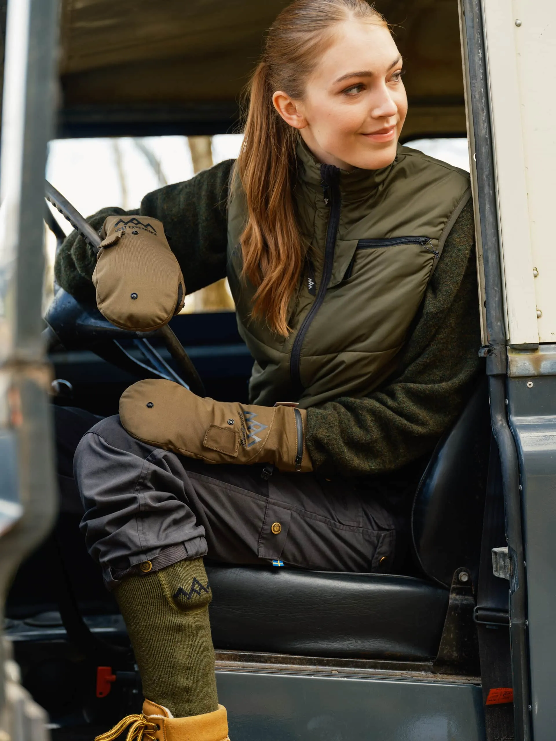 Frau im Auto mit Heizsocken. Grüne Kniestrümpfe, braune Schuhe, olivgrüne Weste, beige Handschuhe.