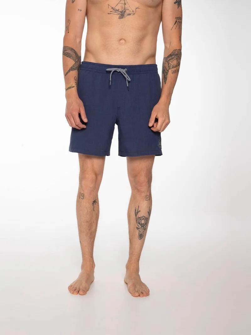 Mann trägt blaue Davey Badeshorts mit Kordelzug, stehend vor weißem Hintergrund. Tattoos auf Armen und Beinen.