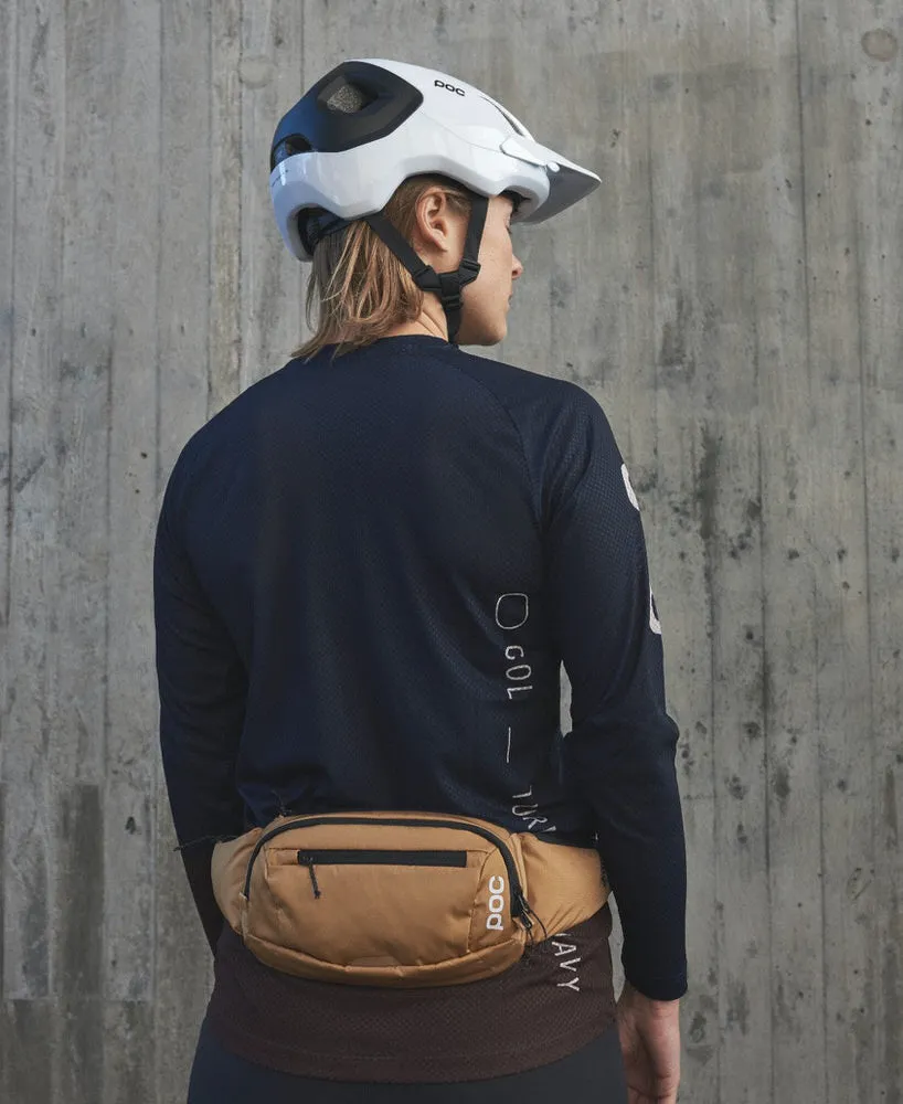 Eine Person trägt eine braune POC Lamina Hüfttasche vor einer Betonwand. Sie trägt auch einen Helm und ein langärmeliges Shirt.