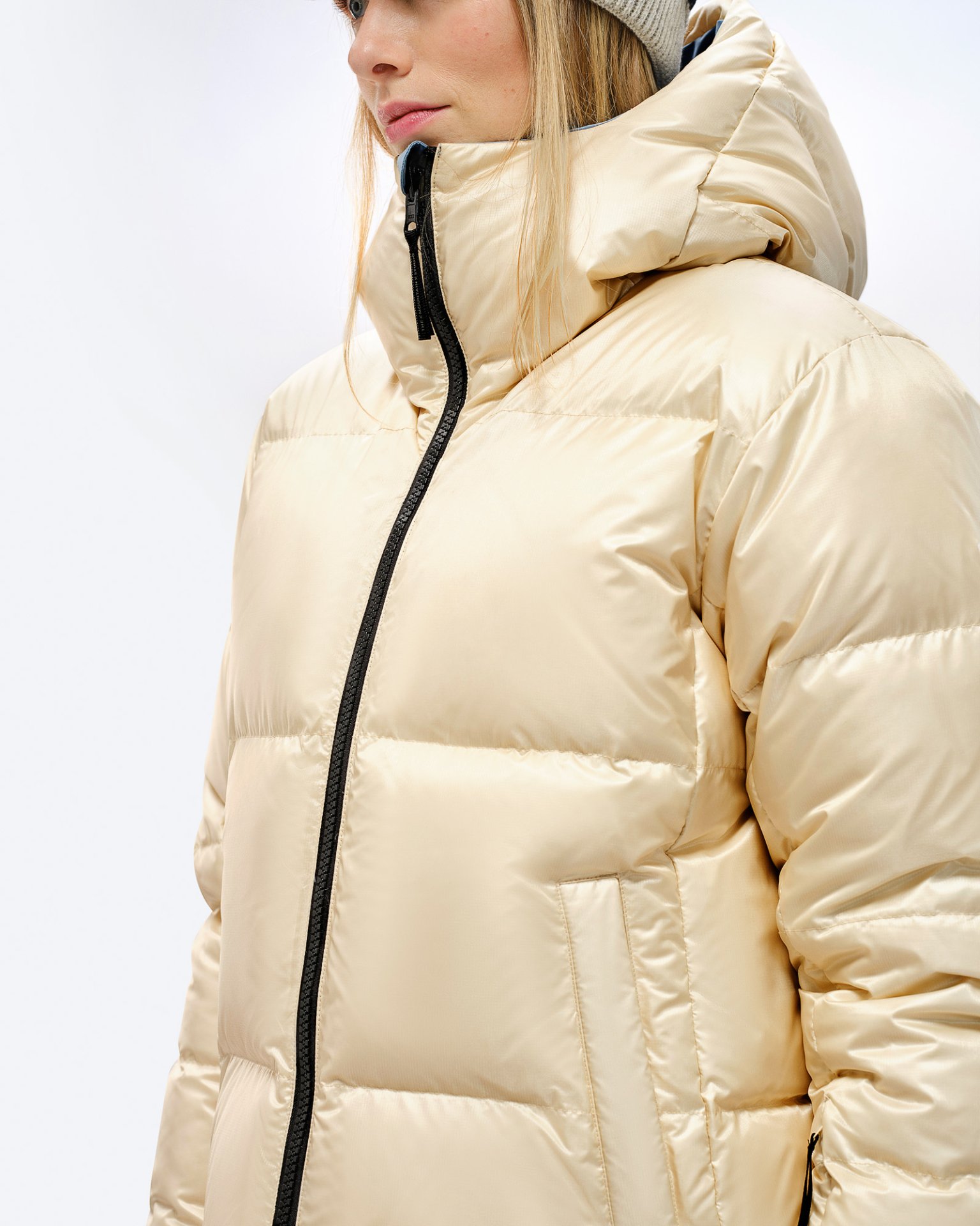 Person in glänzender, beiger Daunenjacke mit Kapuze und schwarzem Reißverschluss. Steppjacke, seitliche Ansicht.